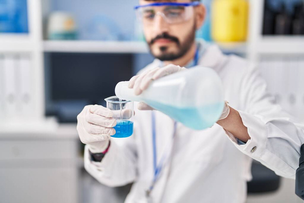 Young hispanic man scientist pouring liquid on glass at laboratory - Фото, изображение