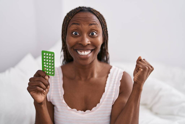 Doğum kontrol hapları tutan Afrikalı Amerikalı kadın gururla bağırıyor, zaferi kutluyor ve başarıyı kollarını kaldırarak kutluyor.  - Fotoğraf, Görsel