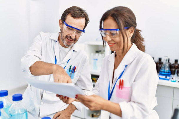 Middle age man and woman partners wearing scientist uniform reading document at laboratory - Photo, Image