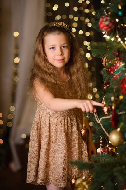 happy girl decorates a christmas tree in the room, new year garland lamps give yellow bokeh on the background - Fotoğraf, Görsel