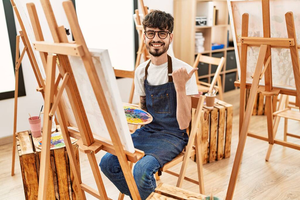 Spanischer Mann mit Bart im Kunststudio zeigt Daumen zur Seite und lächelt glücklich mit offenem Mund  - Foto, Bild