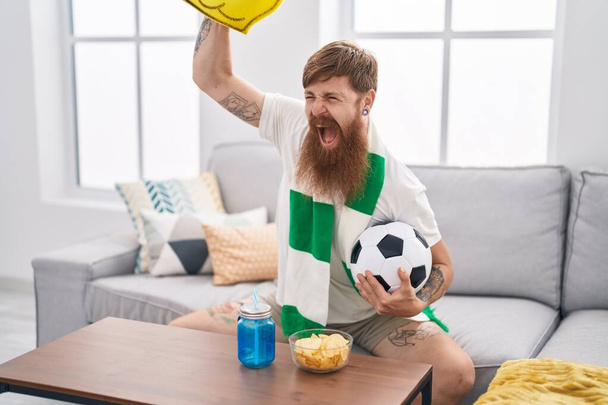Junger Rotschopf unterstützt Fußballmannschaft zu Hause auf dem Sofa - Foto, Bild