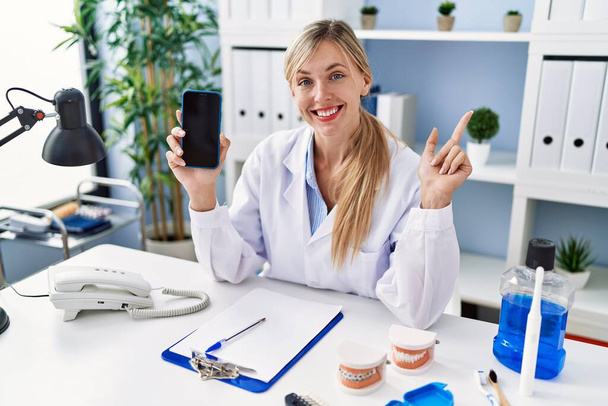 Piękny dentysta kobieta pokazuje ekran smartfona uśmiechnięty szczęśliwy wskazując ręką i palcem w bok  - Zdjęcie, obraz