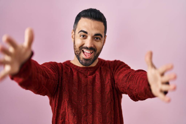 Νεαρός Ισπανός άνδρας με γενειάδα που φοράει casual πουλόβερ πάνω από ροζ φόντο κοιτάζοντας την κάμερα χαμογελώντας με ανοιχτές αγκάλες για αγκαλιά. χαρούμενη έκφραση που αγκαλιάζει την ευτυχία.  - Φωτογραφία, εικόνα