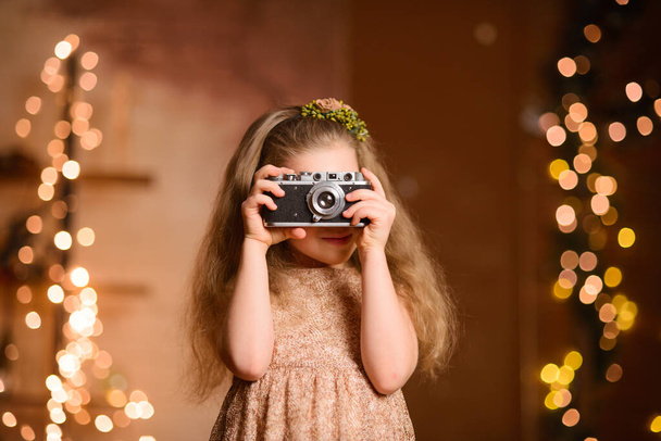 portrait of a little girl in a festive dress with flowing hair takes pictures on a vintage camera. New Year's garland lamps give yellow bokeh on a brown background in the room - Foto, Bild