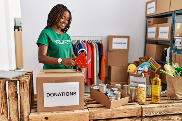 Young african american woman wearing volunteer uniform packing donations box at charity center - Photo, Image
