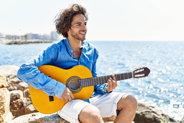 Genç İspanyol adam klasik gitar çalıyor plajda rock 'ın üzerinde oturuyor.. - Fotoğraf, Görsel