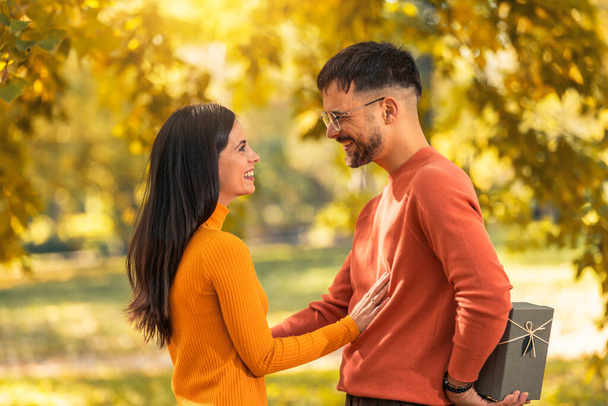 白人のボーイフレンドは、愛の愛情のバレンタインデーの誕生日のシンボルのために存在する秋の公園の驚きで美しいガールフレンドにボックスギフトを与えます - 写真・画像
