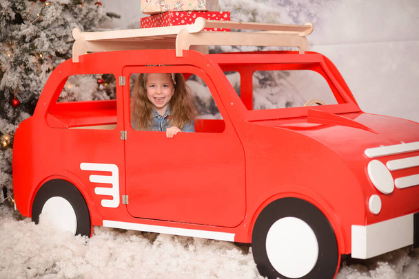 boldog lány ül egy piros játék fa autó, néz ki az ablakon, fehér dekoratív hó között - Fotó, kép