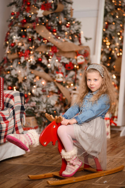Ευτυχισμένο κορίτσι κάθεται σε ένα κόκκινο παιχνίδι ξύλινο κουνιστό άλογο, σε ένα δωμάτιο με ένα χριστουγεννιάτικο δέντρο, ξύλινο πάτωμα. κίτρινα φώτα γιρλάντες αντανακλώνται στον καθρέφτη - Φωτογραφία, εικόνα