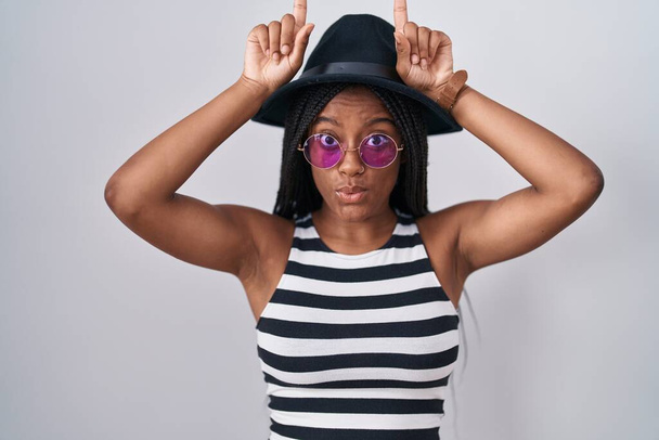 Молодий афроамериканський американець з косами в капелюсі і сонцезахисних окулярах робить смішний жест з пальцем над головою як бульбашкові роги
  - Фото, зображення