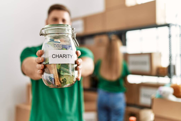 Ein junges hispanisches Freiwilligenpaar, das in einem Wohltätigkeitszentrum arbeitet. Mann lächelt glücklich und hält Spendendose mit Geld in der Hand. - Foto, Bild