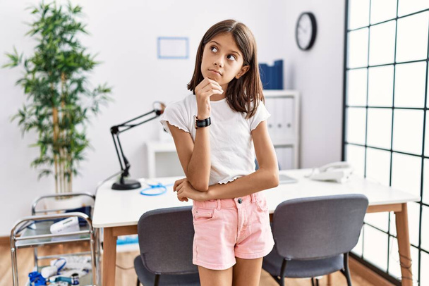 Young hispanic girl standing at pediatrician clinic with hand on chin thinking about question, pensive expression. smiling with thoughtful face. doubt concept.  - Photo, Image