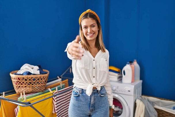 Mujer rubia joven en la sala de lavandería sonriendo amable ofreciendo apretón de manos como saludo y bienvenida. negocios exitosos.  - Foto, Imagen