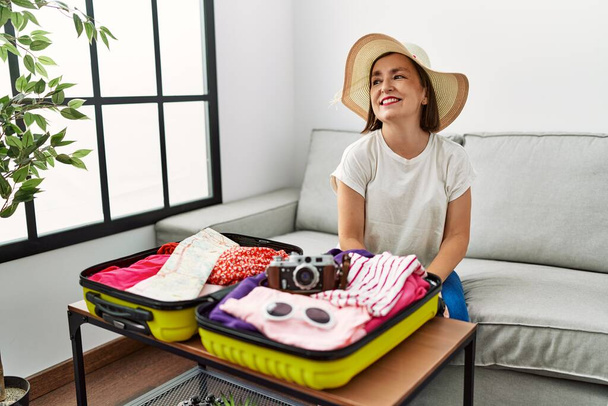 Belle femme hispanique d'âge moyen emballant des vêtements d'été dans une valise regardant de côté avec le sourire sur le visage, expression naturelle. riant confiant.  - Photo, image