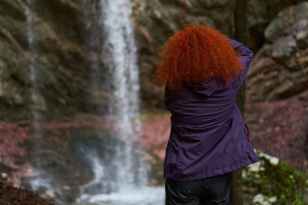 Woman nature photographer with backpack and camera hiking by a waterfall in the mountains - Photo, image