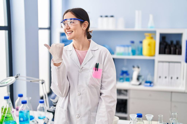 Молодая брюнетка, работающая в научной лаборатории, указывая пальцем вверх в сторону улыбаясь счастливой с открытым ртом  - Фото, изображение