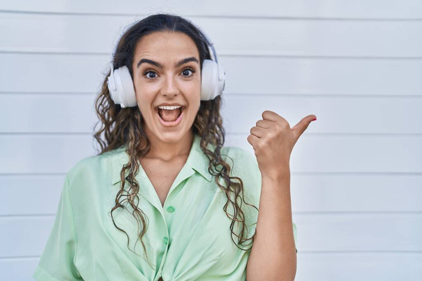 Jovem hispânica ouvindo música usando fones de ouvido apontando o polegar para o lado sorrindo feliz com a boca aberta  - Foto, Imagem