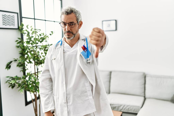 Middle age hispanic man wearing doctor uniform and stethoscope at waiting room looking unhappy and angry showing rejection and negative with thumbs down gesture. bad expression.  - Valokuva, kuva