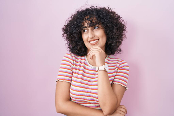 Νεαρή γυναίκα της Μέσης Ανατολής στέκεται πάνω από ροζ φόντο αναζητούν αυτοπεποίθηση στην κάμερα χαμογελώντας με σταυρωμένα χέρια και το χέρι έθεσε στο πηγούνι. θετική σκέψη.  - Φωτογραφία, εικόνα