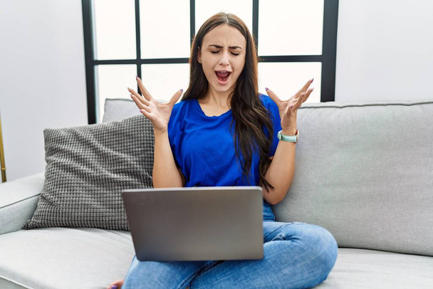 Νεαρή μελαχρινή γυναίκα που χρησιμοποιεί φορητό υπολογιστή στο σπίτι γιορτάζει τρελός και τρελός για την επιτυχία με τα χέρια ψηλά και κλειστά μάτια ουρλιάζοντας ενθουσιασμένος. έννοια νικητή  - Φωτογραφία, εικόνα