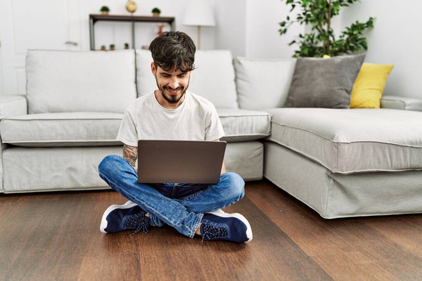 Giovane uomo ispanico utilizzando il computer portatile seduto sul pavimento a casa. - Foto, immagini