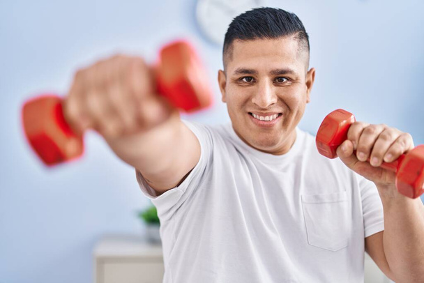 Молодой латинос уверенно улыбается, используя гантели для бокса дома - Фото, изображение