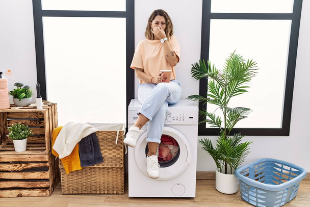 Young hispanic woman drinking coffee waiting for washing machine at laundry room smelling something stinky and disgusting, intolerable smell, holding breath with fingers on nose. bad smell  - Foto, imagen