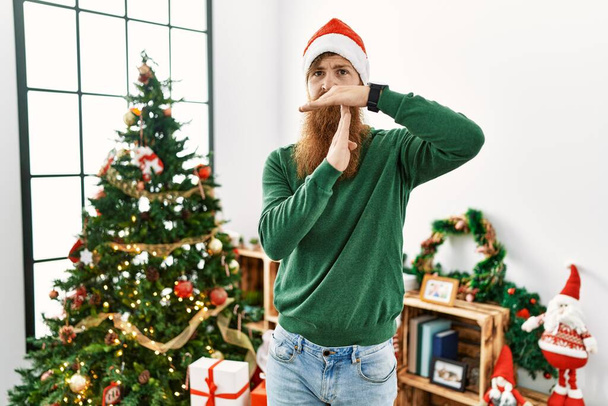 Rotschopf mit langem Bart trägt Weihnachtsmütze am Weihnachtsbaum und macht Auszeit mit Händen, frustriertem und ernstem Gesicht  - Foto, Bild
