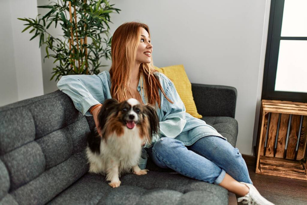 Junge Kaukasierin lächelt selbstbewusst und sitzt zu Hause mit Hund auf dem Sofa - Foto, Bild