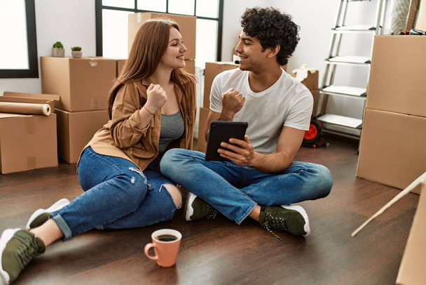 Nuori pari hymyilee onnellisena käyttämällä kosketuslevyä ja juomalla kahvia uudessa kodissa. - Valokuva, kuva