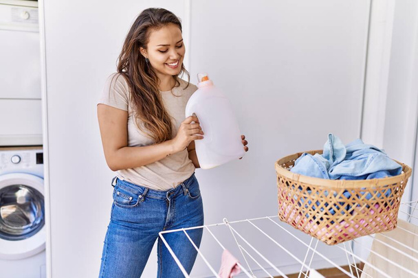 Young hispanic girl doing laundry holding detergent bottle at home - Foto, imagen