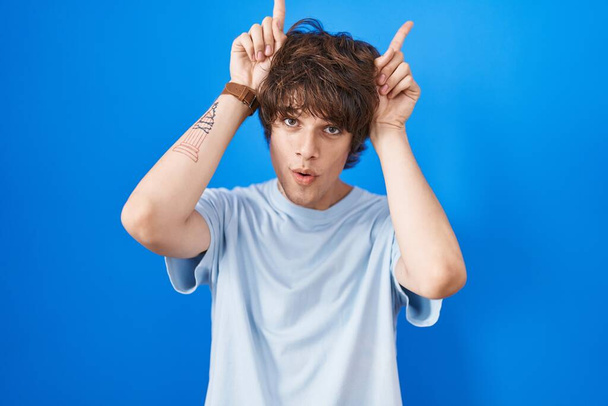 Spaanse jongeman die over een blauwe achtergrond staat en grappig gebaar maakt met vinger over het hoofd als stierenhoorns  - Foto, afbeelding