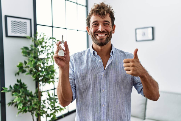 Молодой красивый мужчина держит ключи от нового дома улыбаясь счастливым и позитивным, большой палец вверх делает отлично и знак одобрения  - Фото, изображение