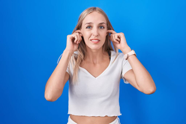 Junge kaukasische Frau, die vor blauem Hintergrund steht und die Ohren mit genervten Fingern vor lauter Musik verdeckt. Gehörloses Konzept.  - Foto, Bild