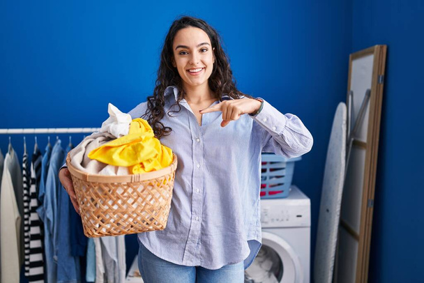 Young brunette woman holding laundry basket pointing finger to one self smiling happy and proud  - Photo, Image