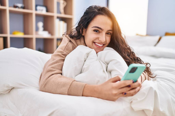 Young hispanic woman using smartphone lying on bed at bedroom - Foto, imagen