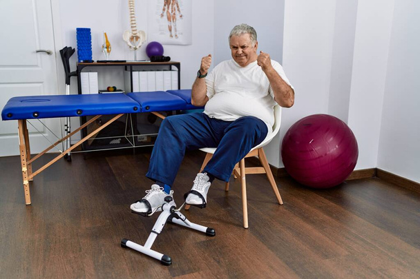 Старший кавказький чоловік у фізіотерапевтичній клініці, використовуючи педальні вправи, збуджений успіхом з піднятими руками і закритими очима, святкуючи перемогу посміхаючись. концепція переможця
.  - Фото, зображення