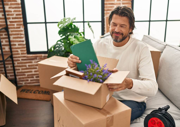 Hombre de mediana edad sonriendo confiado desembalaje caja de cartón en el nuevo hogar - Foto, Imagen