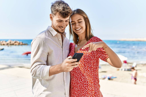 Молодая латиноамериканская пара в отпуске улыбается счастливо, используя смартфон на пляже - Фото, изображение