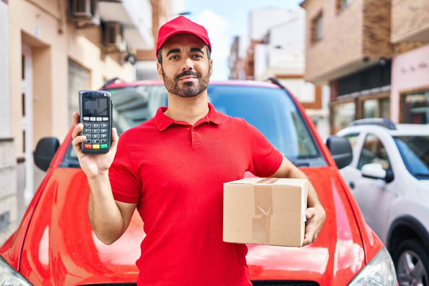 Young hispanic man with beard wearing delivery uniform and cap holding dataphone relaxed with serious expression on face. simple and natural looking at the camera.  - Foto, afbeelding