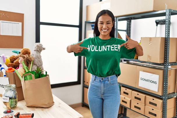 jonge latijn vrouw wijzend met vingers naar vrijwilliger uniform op liefdadigheidscentrum - Foto, afbeelding