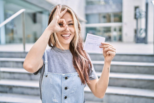 Joven mujer caucásica sosteniendo tarjeta de registro covid sonriendo feliz haciendo signo de ok con la mano en el ojo mirando a través de los dedos  - Foto, imagen