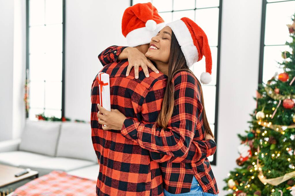 Młoda Latynoska para uśmiechnięta szczęśliwa i przytulona trzymająca świąteczny prezent w domu. - Zdjęcie, obraz