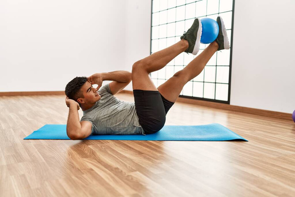 Young hispanic man training abs exercise using ball at sport center - Foto, Imagen