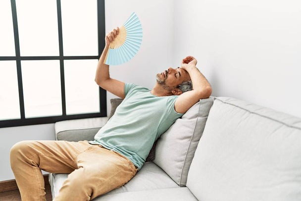 Седовласый мужчина средних лет с помощью ручного вентилятора сидит дома на диване - Фото, изображение