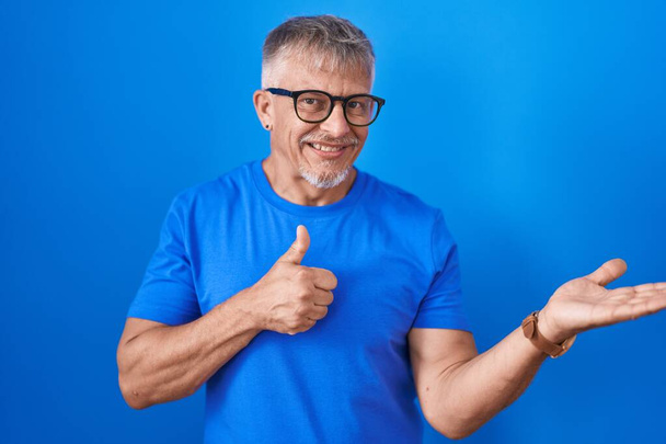 Hombre hispano con el pelo gris de pie sobre fondo azul mostrando la palma de la mano y haciendo buen gesto con los pulgares hacia arriba, sonriendo feliz y alegre  - Foto, Imagen