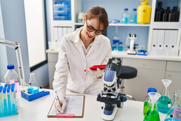 Laboratuvarda çalışan dokunmatik ped kullanan, bilim adamı üniforması giyen beyaz bir kadın. - Fotoğraf, Görsel