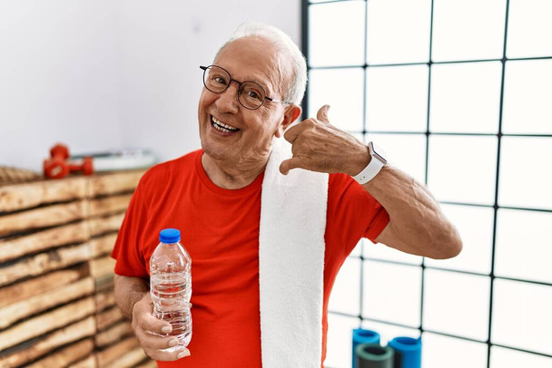 Homme âgé portant des vêtements de sport et serviette à la salle de gym souriant faire geste de téléphone avec la main et les doigts comme parler au téléphone. communiquer les concepts.  - Photo, image