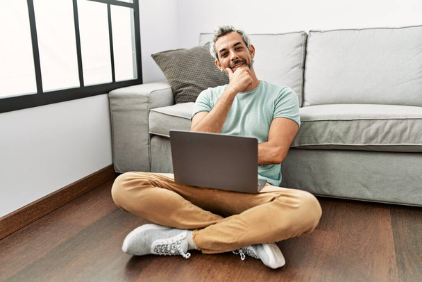 Hispanischer Mann mittleren Alters sitzt mit Laptop auf dem Boden im Wohnzimmer und blickt selbstbewusst in die Kamera, mit verschränkten Armen und erhobener Hand am Kinn. Positives Denken.  - Foto, Bild